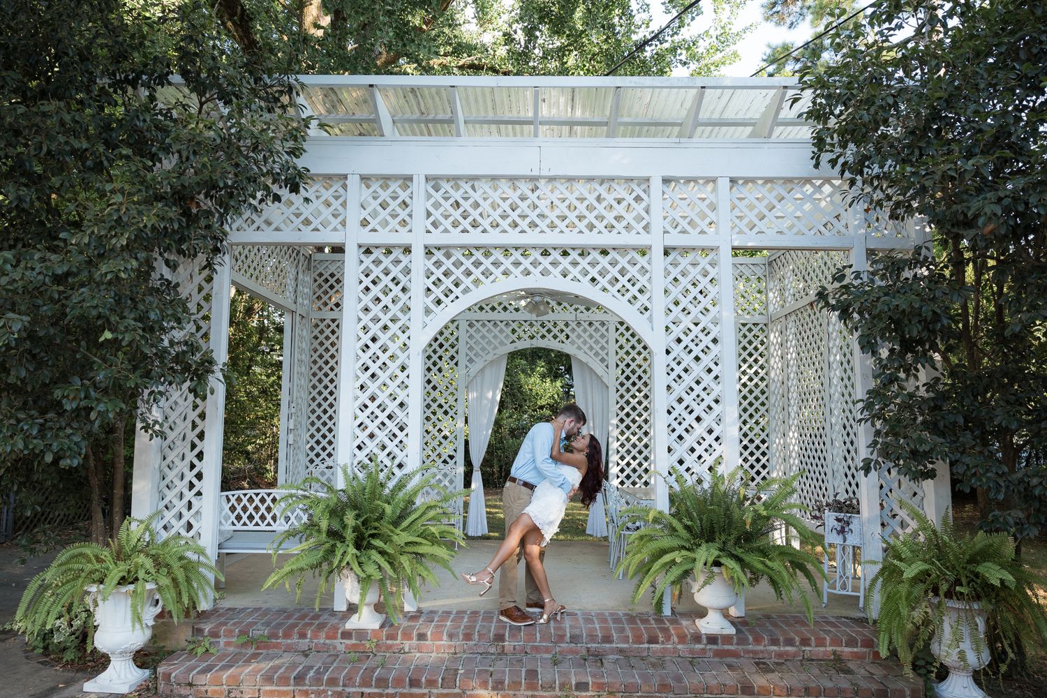 Couple sharing a romantic kiss under a white lattice garden pergola surrounded by lush ferns and trees.