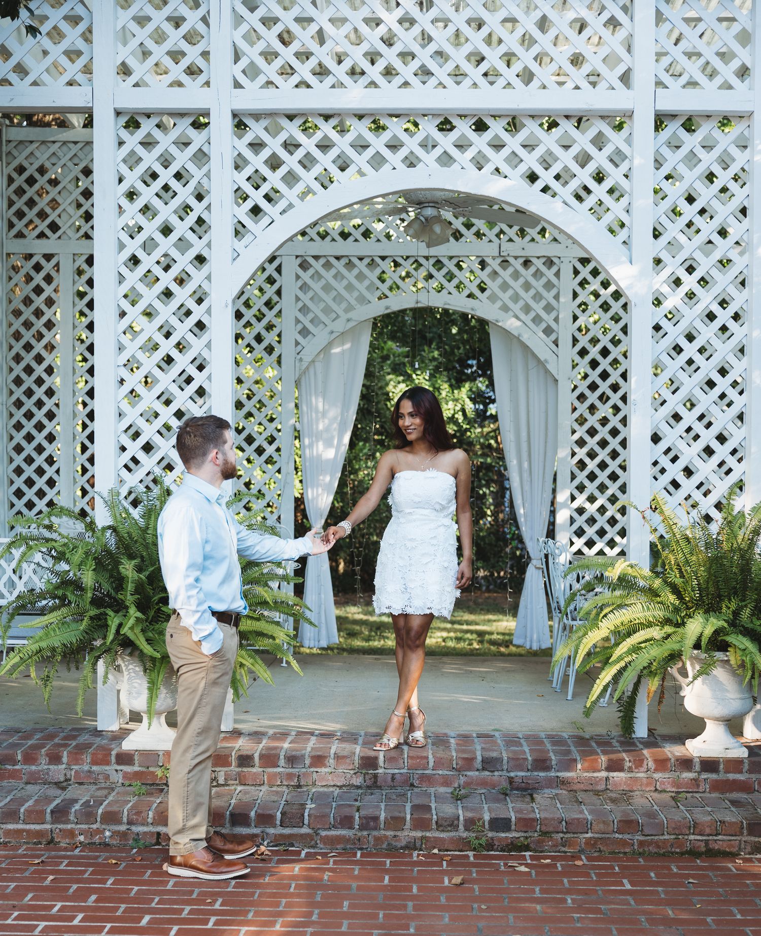 A couple holds hands under a white lattice gazebo arch surrounded by ferns on brick.