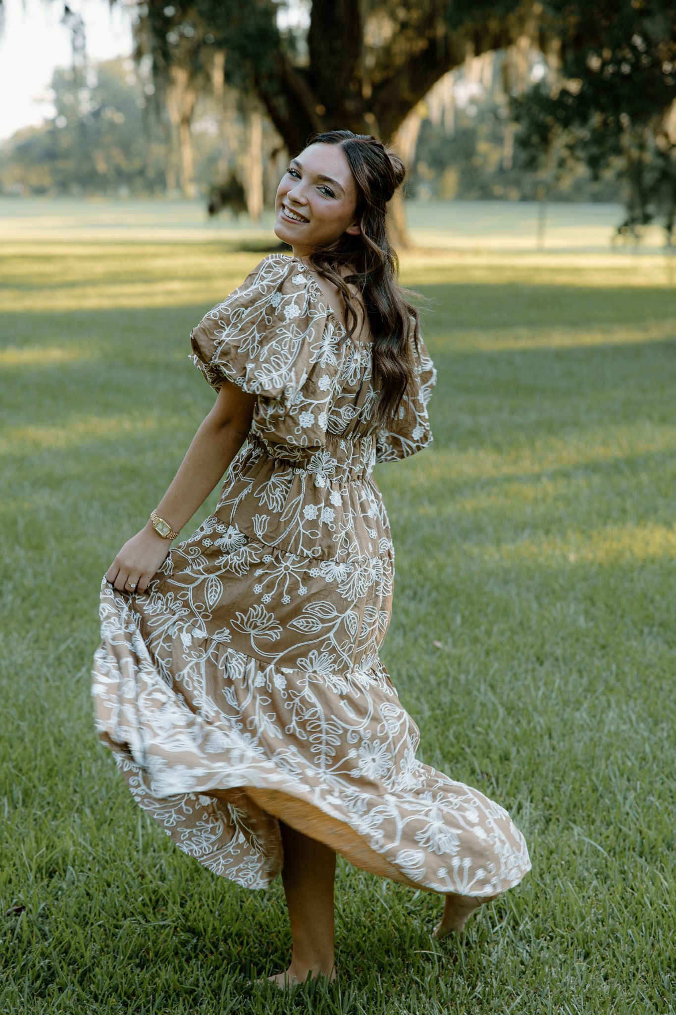 Woman in floral boho maxi dress twirling on green grass with Spanish moss trees in golden hour light.