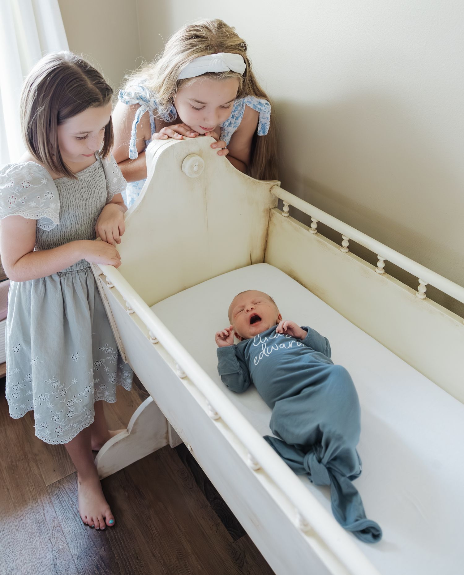 Two young girls in dresses lean over a white crib to look at their yawning newborn sibling in a teal sleeper.