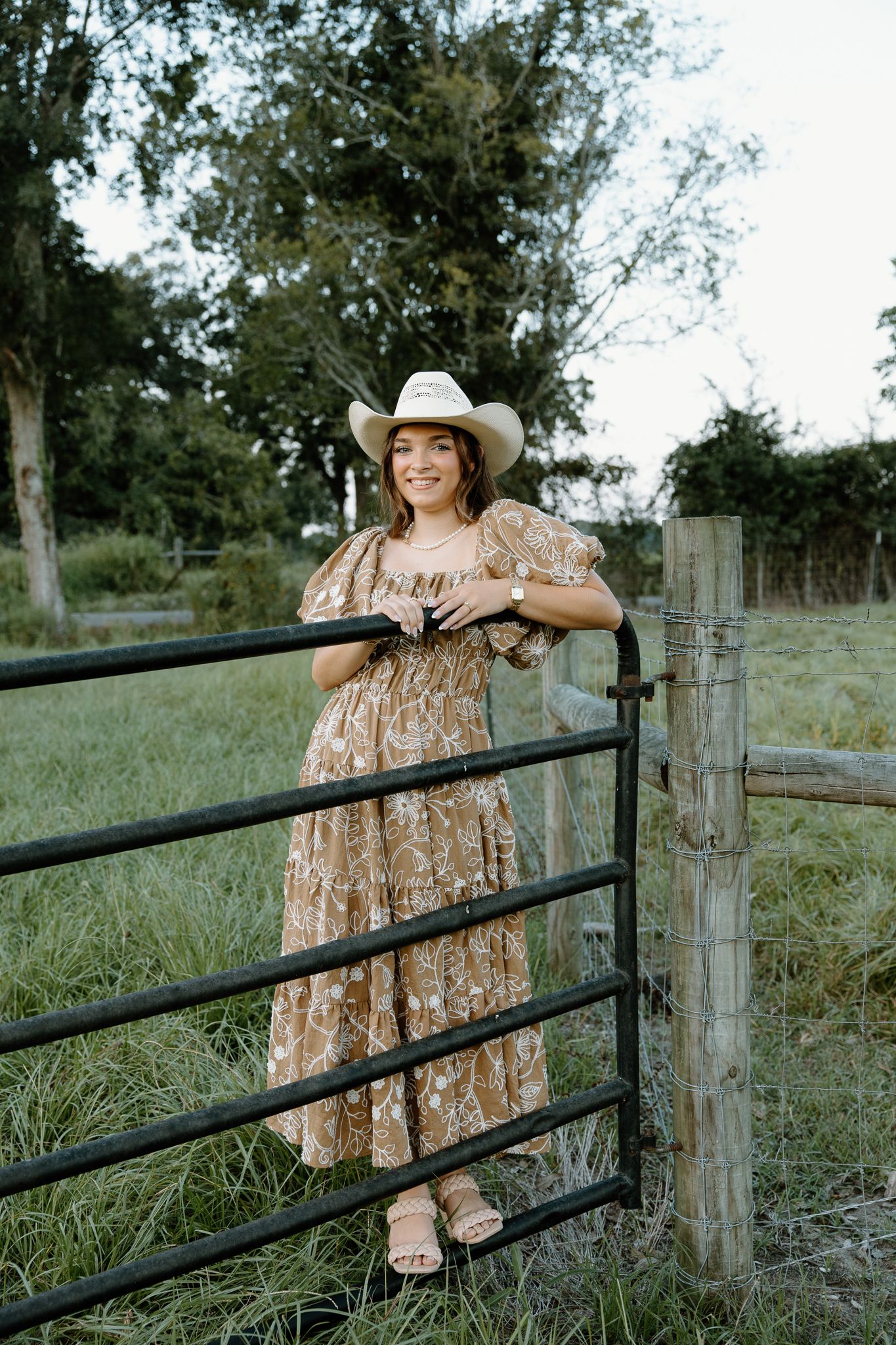 Woman in floral maxi dress and white cowboy hat leaning on black farm gate with green pasture background.
