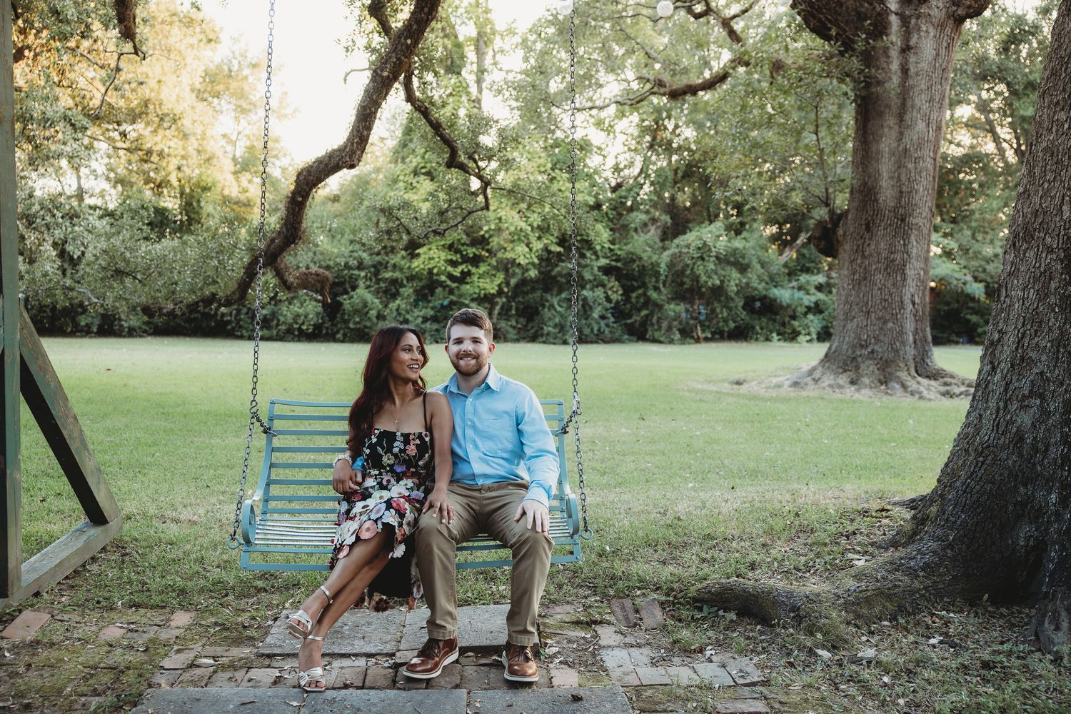 Couple sitting together on a light blue wooden swing in a lush green park, smiling and posing for an engagement photo.