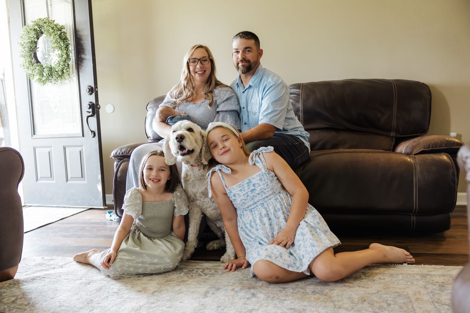 Candid family photo with parents, two girls, white dog, and newborn baby on living room floor.