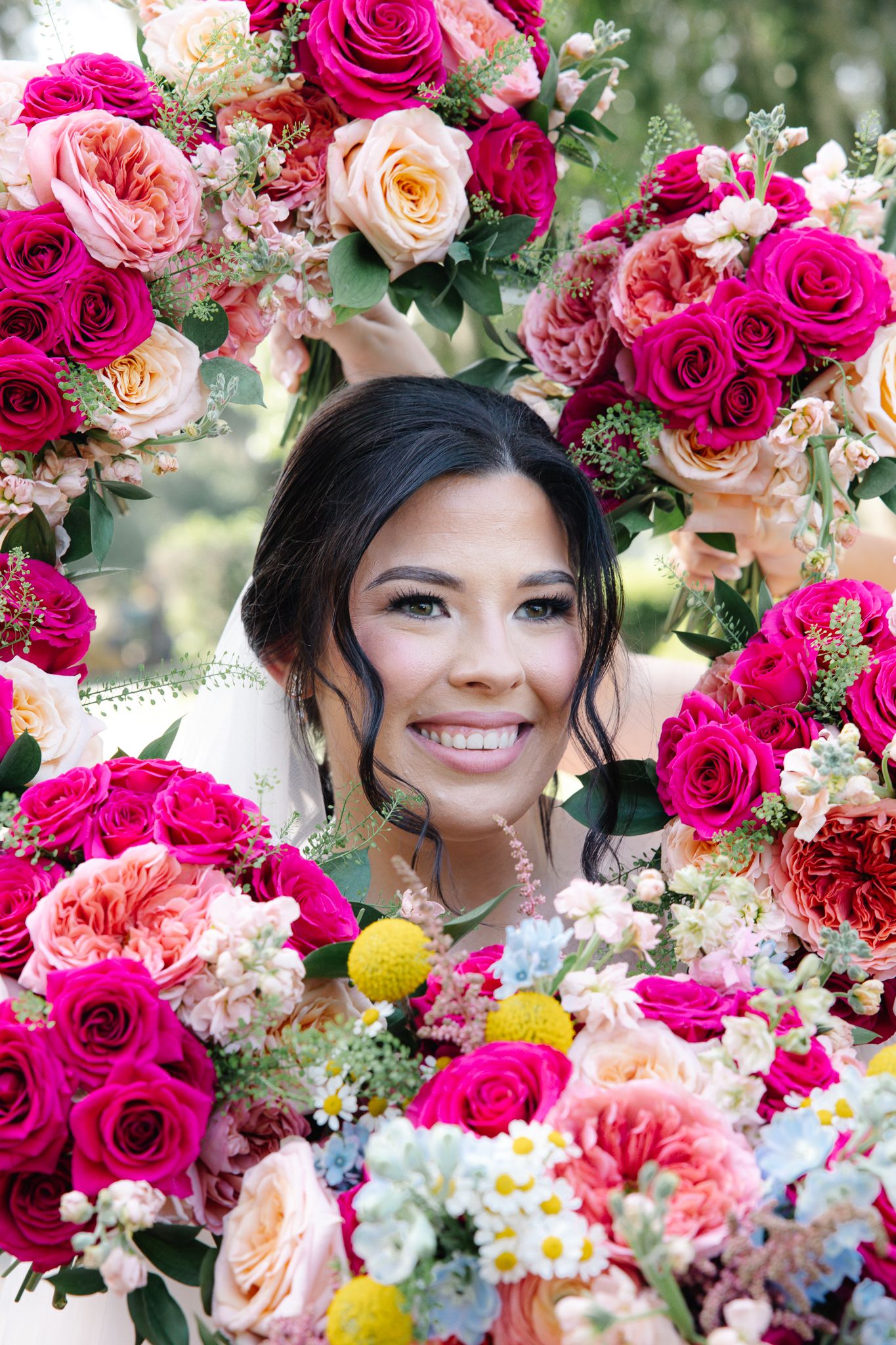 Smiling bride peeks through a stunning circular arrangement of vibrant pink, peach, and white roses.
