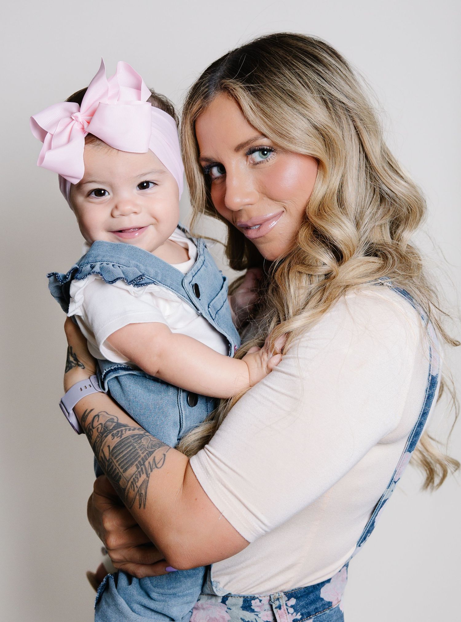 Blonde mother with tattoos holding smiling baby girl wearing pink bow headband and denim outfit.