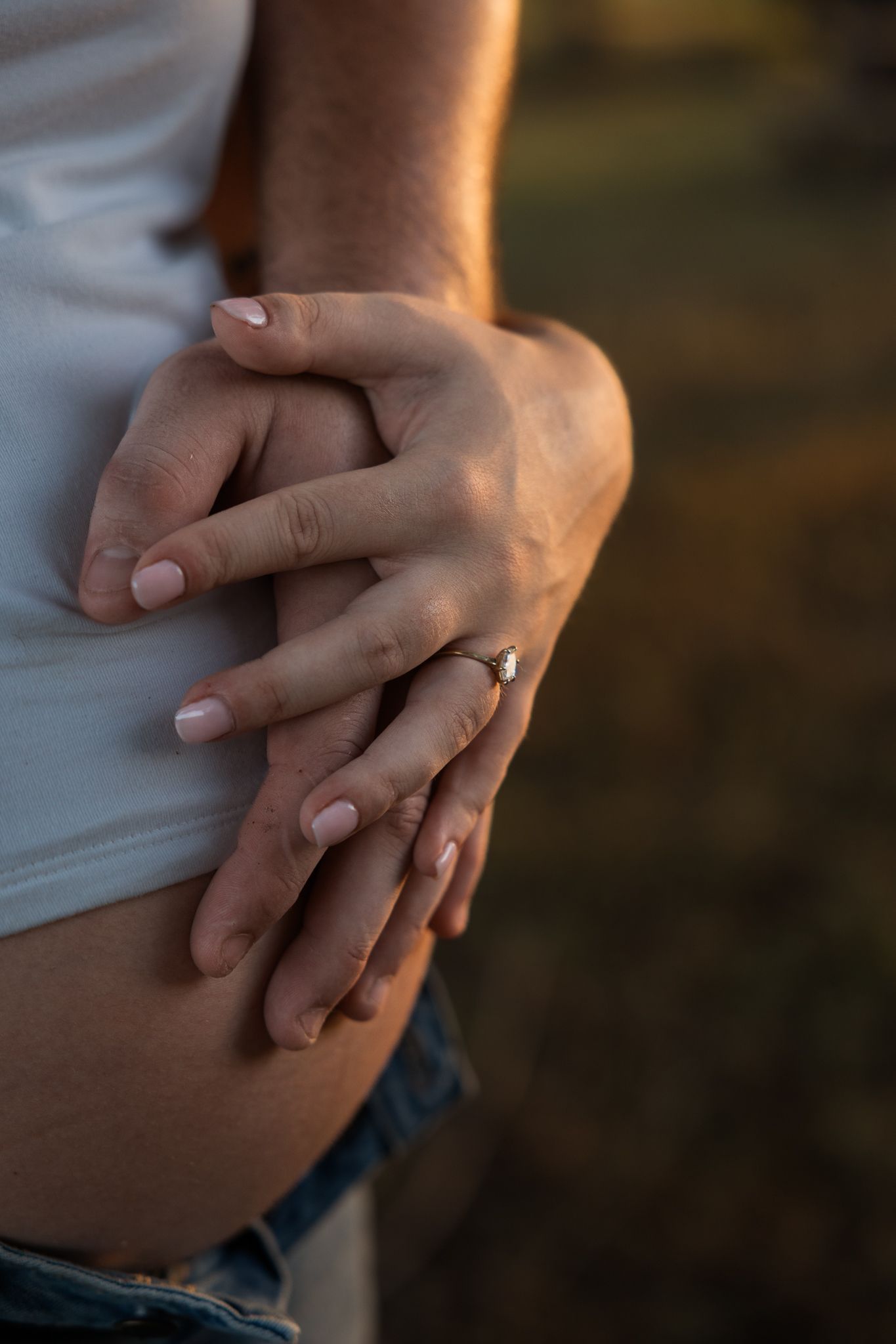 Close-up of couple holding hands on pregnant belly, woman wearing engagement ring, golden sunset light.