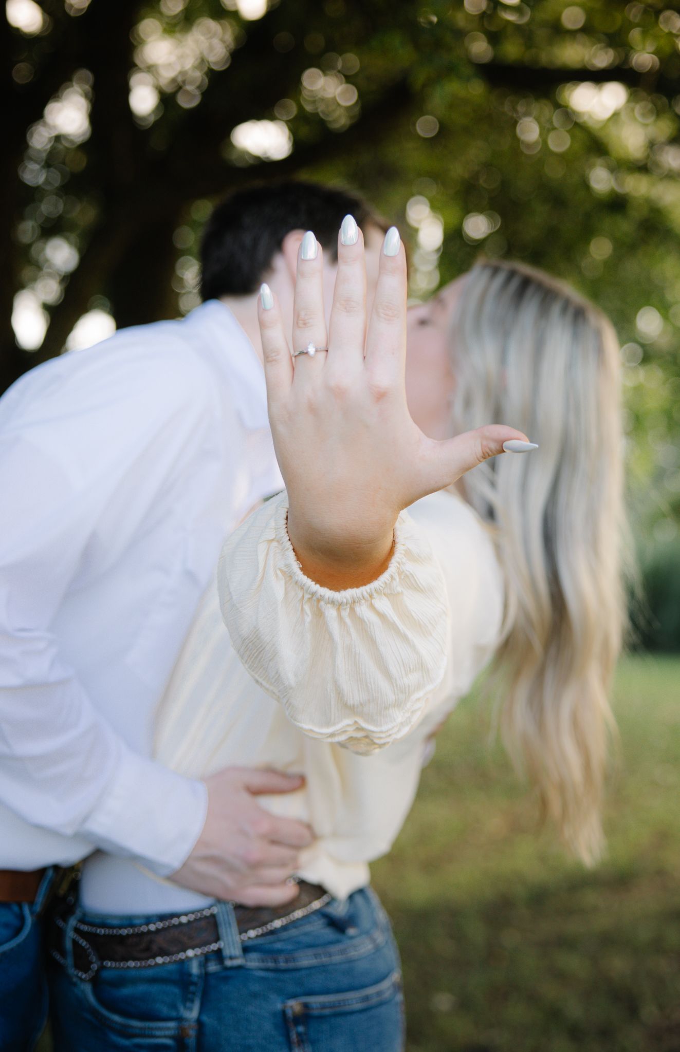 Engaged couple kissing outdoors while woman displays diamond engagement ring toward camera.