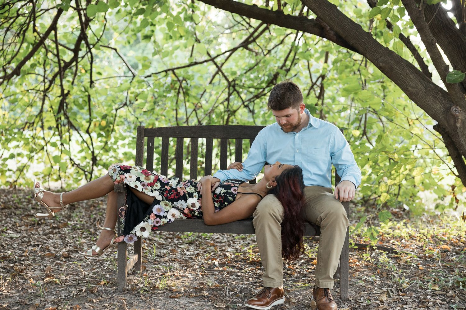 Couple relaxing on a wooden bench outdoors, woman lying across man's lap in a floral dress amid lush green trees.