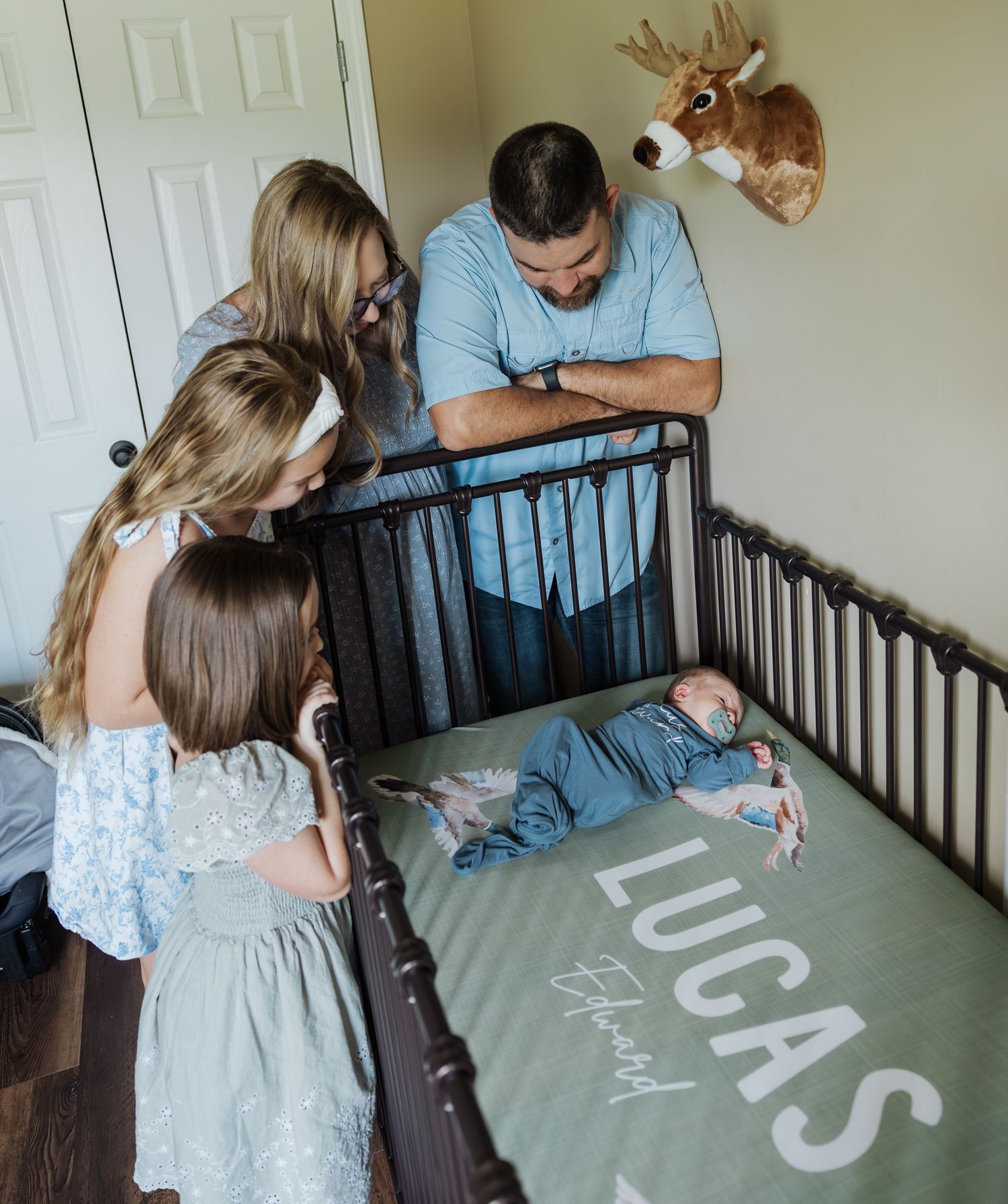 Family of four lovingly gazing at newborn baby Lucas Edward in his crib with personalized blanket.
