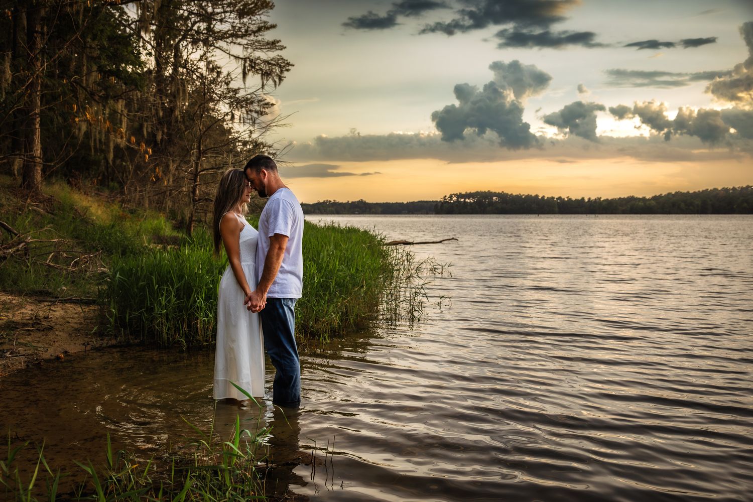 Couple standing in lake water at sunset, holding hands near lush green shoreline with dramatic cloudy sky.