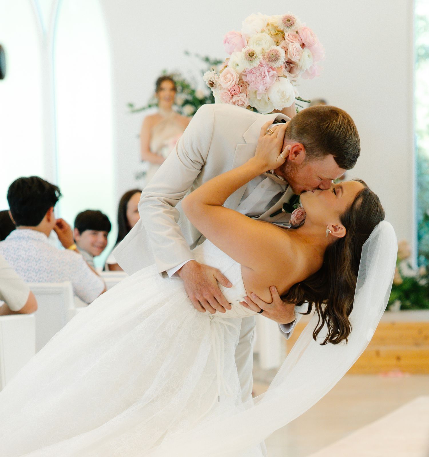 Groom dipping bride for a romantic kiss while holding pink bouquet during indoor wedding reception.