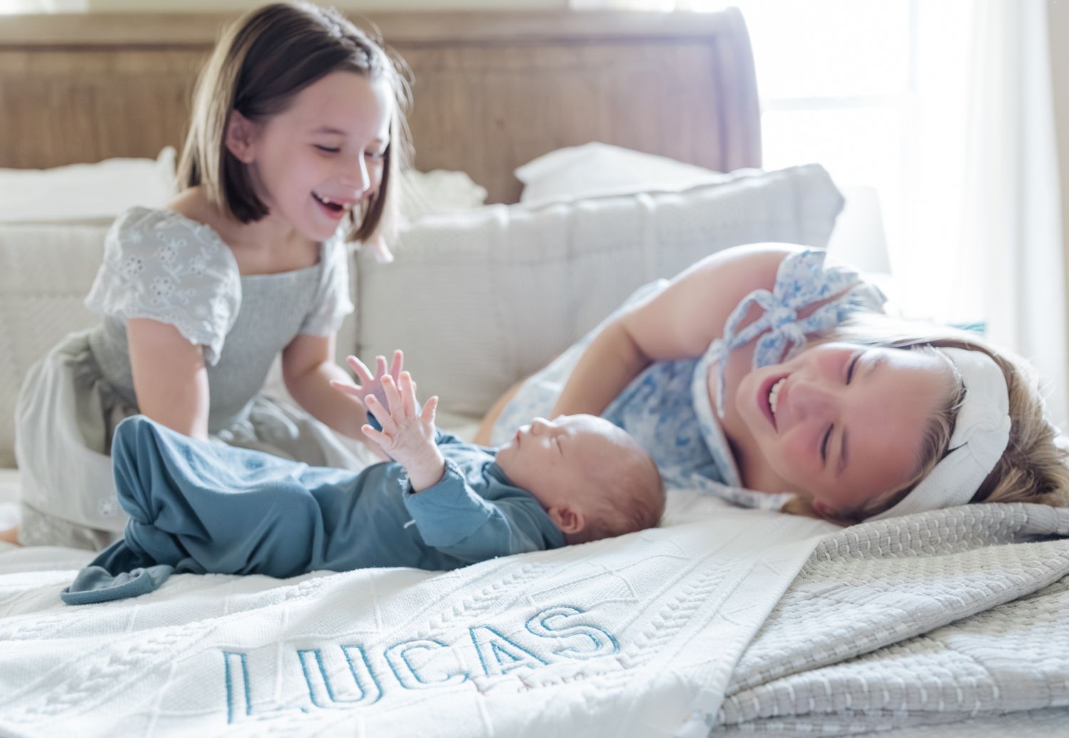 Two girls laugh and play with newborn baby Lucas on a personalized white blanket on a sunlit bed.