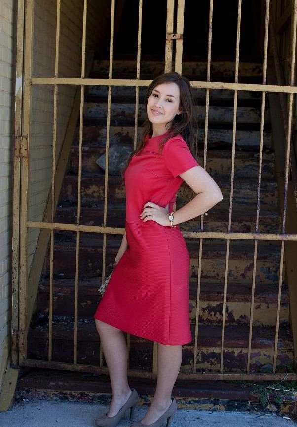 Young woman in fitted red midi dress posing confidently by rustic iron gate outdoors.