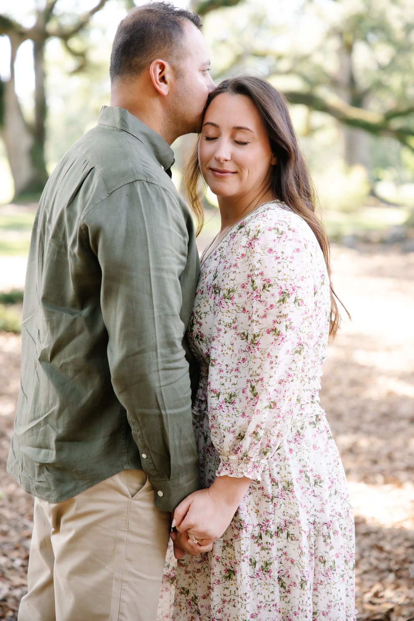 Man in green shirt kissing woman in floral dress on forehead while holding hands outdoors.