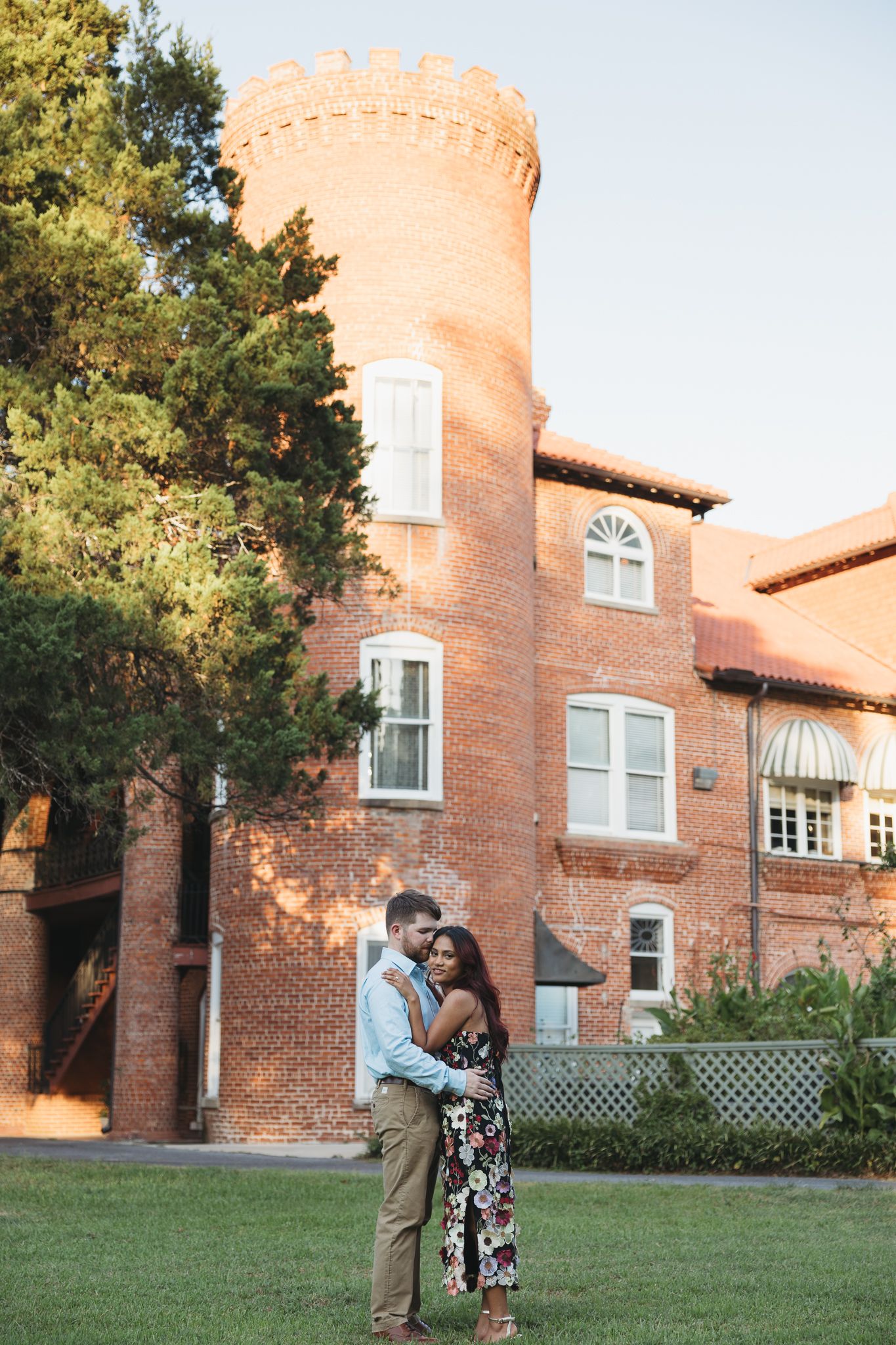 Couple embracing in front of a historic red brick building with a castle tower at golden hour.