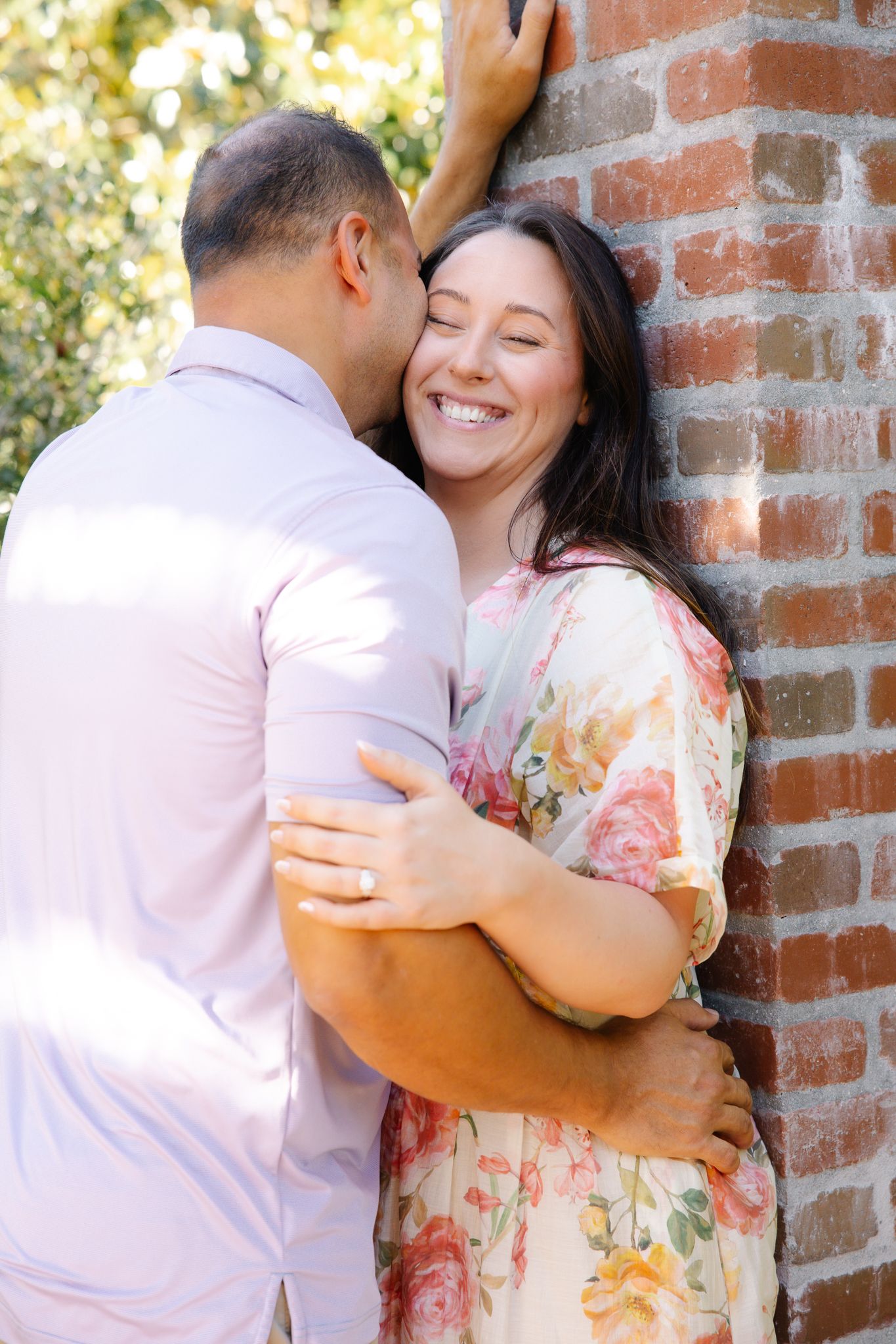 Woman smiling joyfully as man embraces her against a rustic brick wall outdoors.