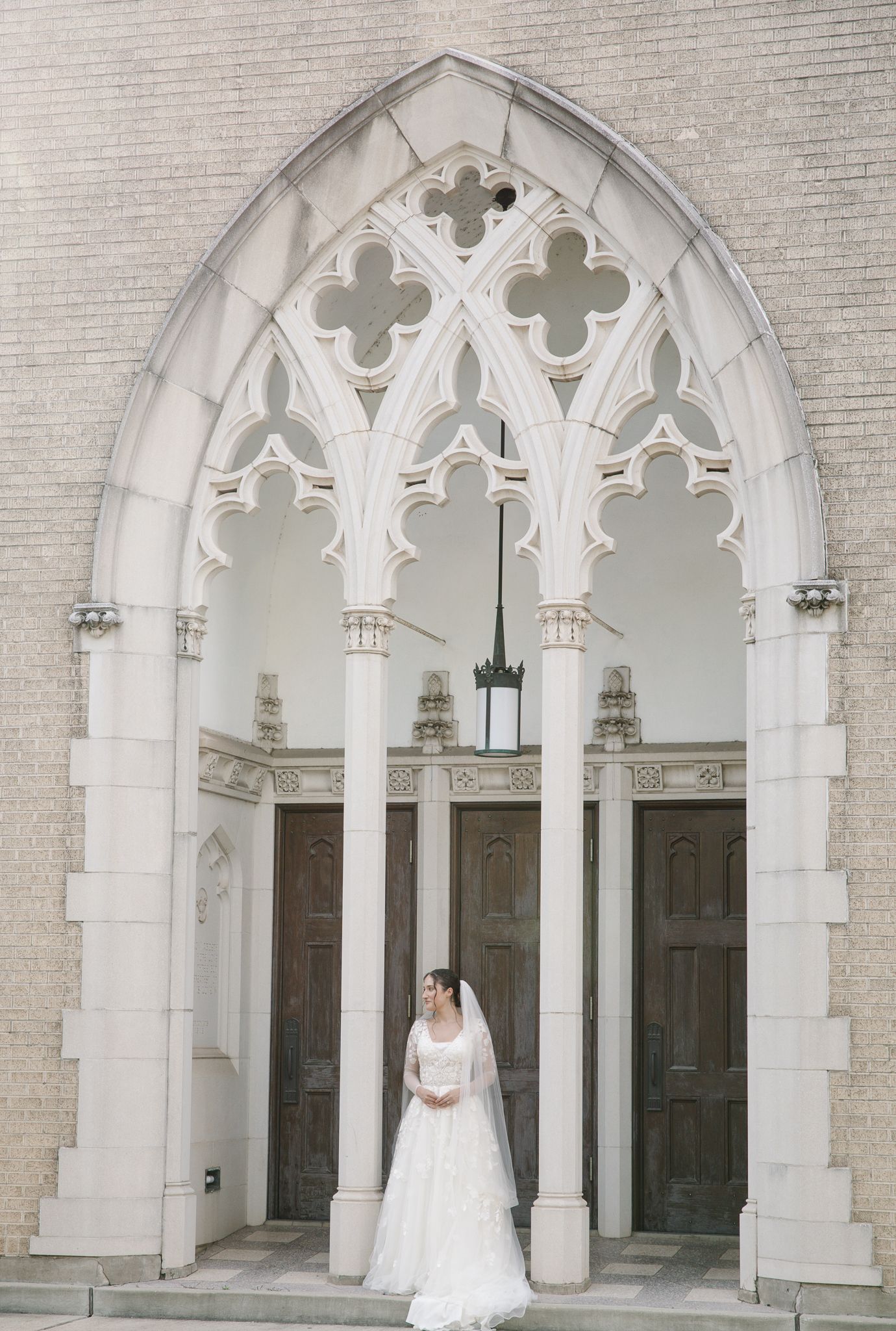Bride in a white lace gown and veil stands beneath a stunning Gothic church archway with ornate stone columns.