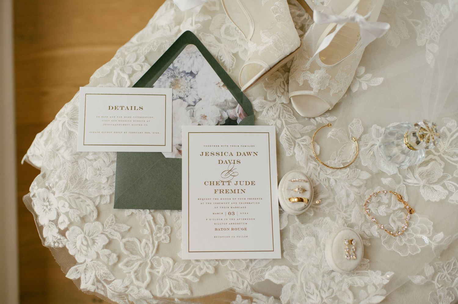 Wedding flat lay with green-accented invitation suite, bridal shoes, rings, and floral details on lace gown.