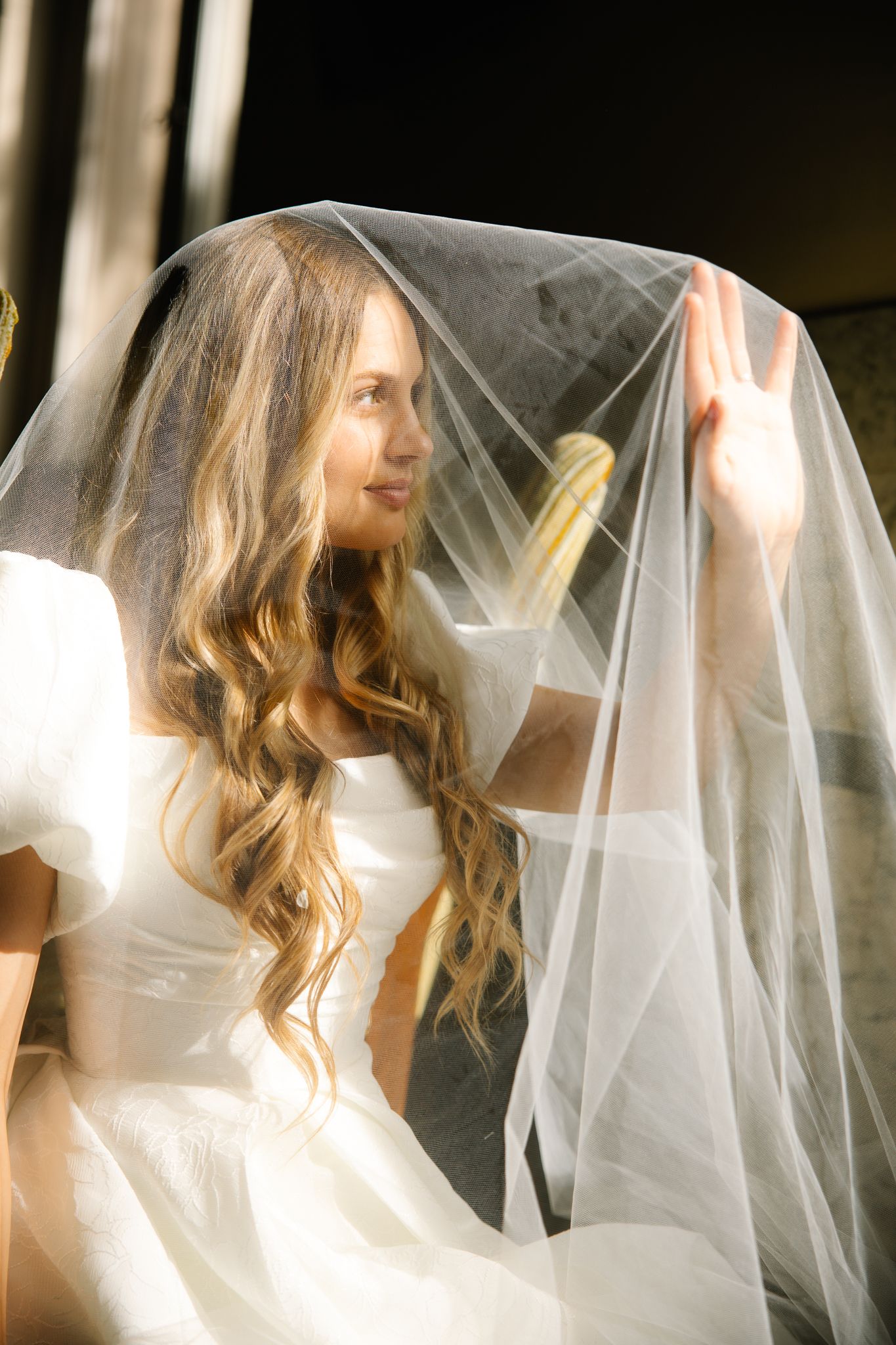 Bride lifting her sheer wedding veil with long wavy hair wearing a white bridal gown in soft natural light.