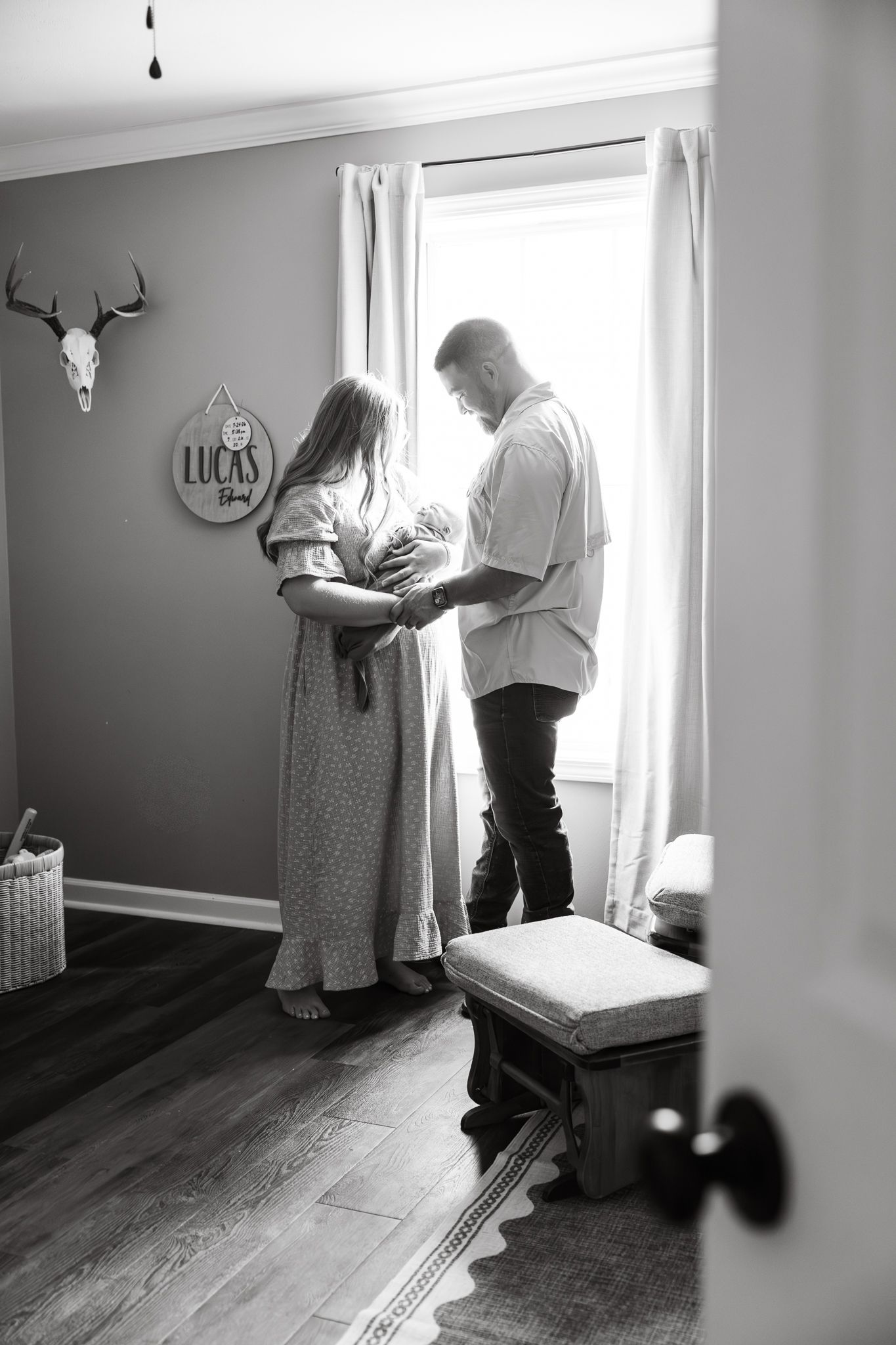 Black and white photo of parents standing by a window holding their newborn in a nursery with a Lucas sign.