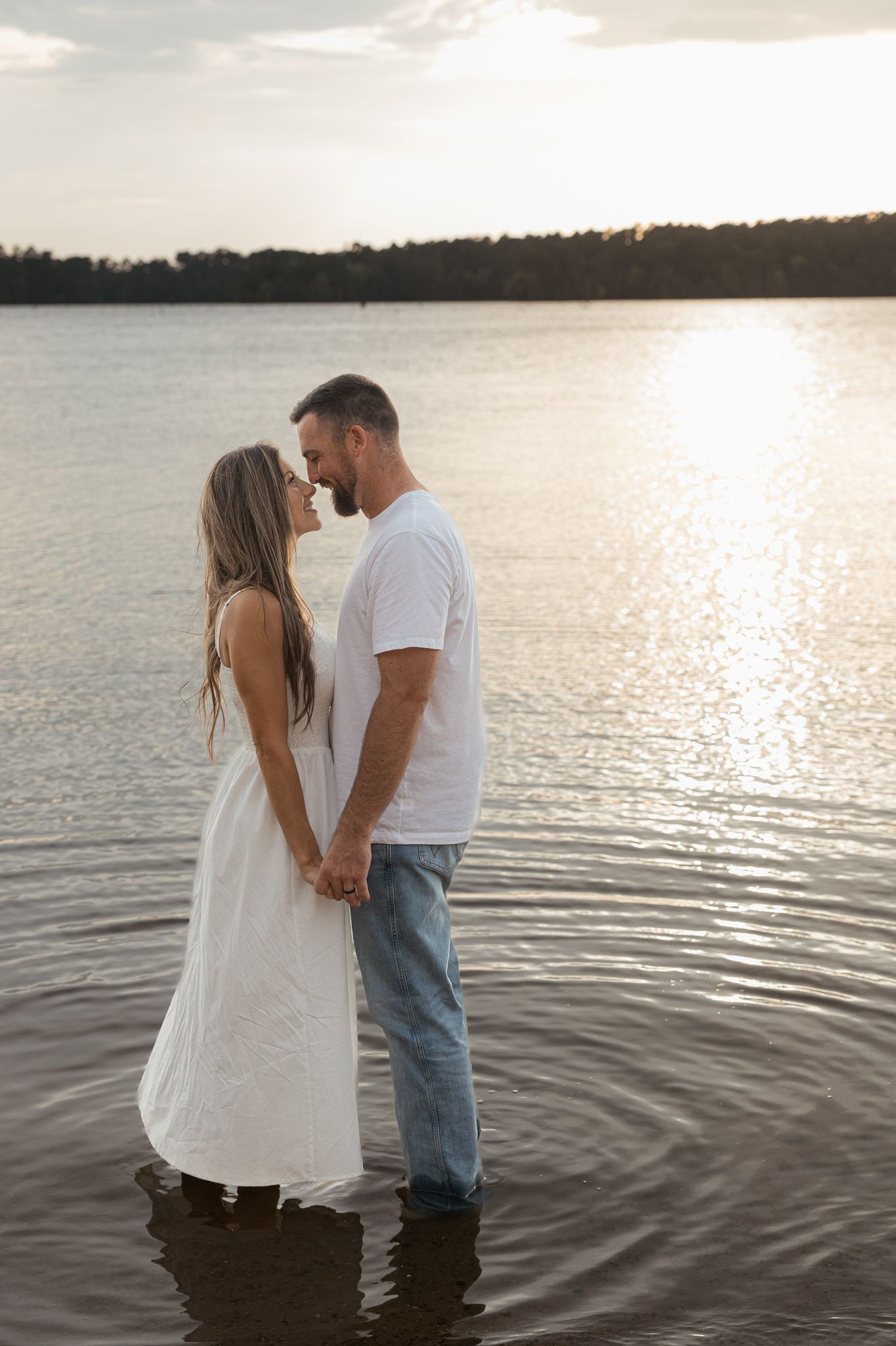 Couple standing in lake water holding hands at sunset, woman in white dress, man in jeans and white tee.