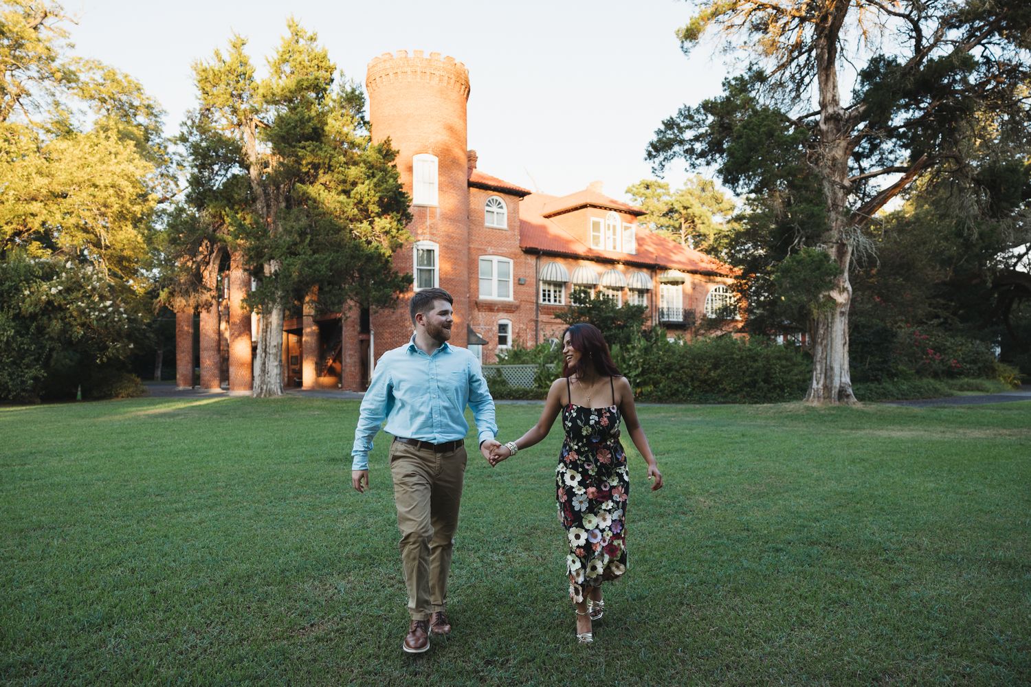 Couple holding hands walking across green lawn in front of historic red brick castle building at golden hour.