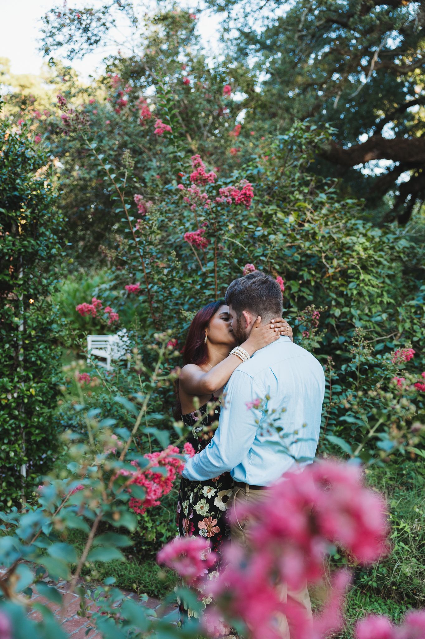 Couple sharing a romantic kiss surrounded by lush green bushes and vibrant pink flowers in a garden.