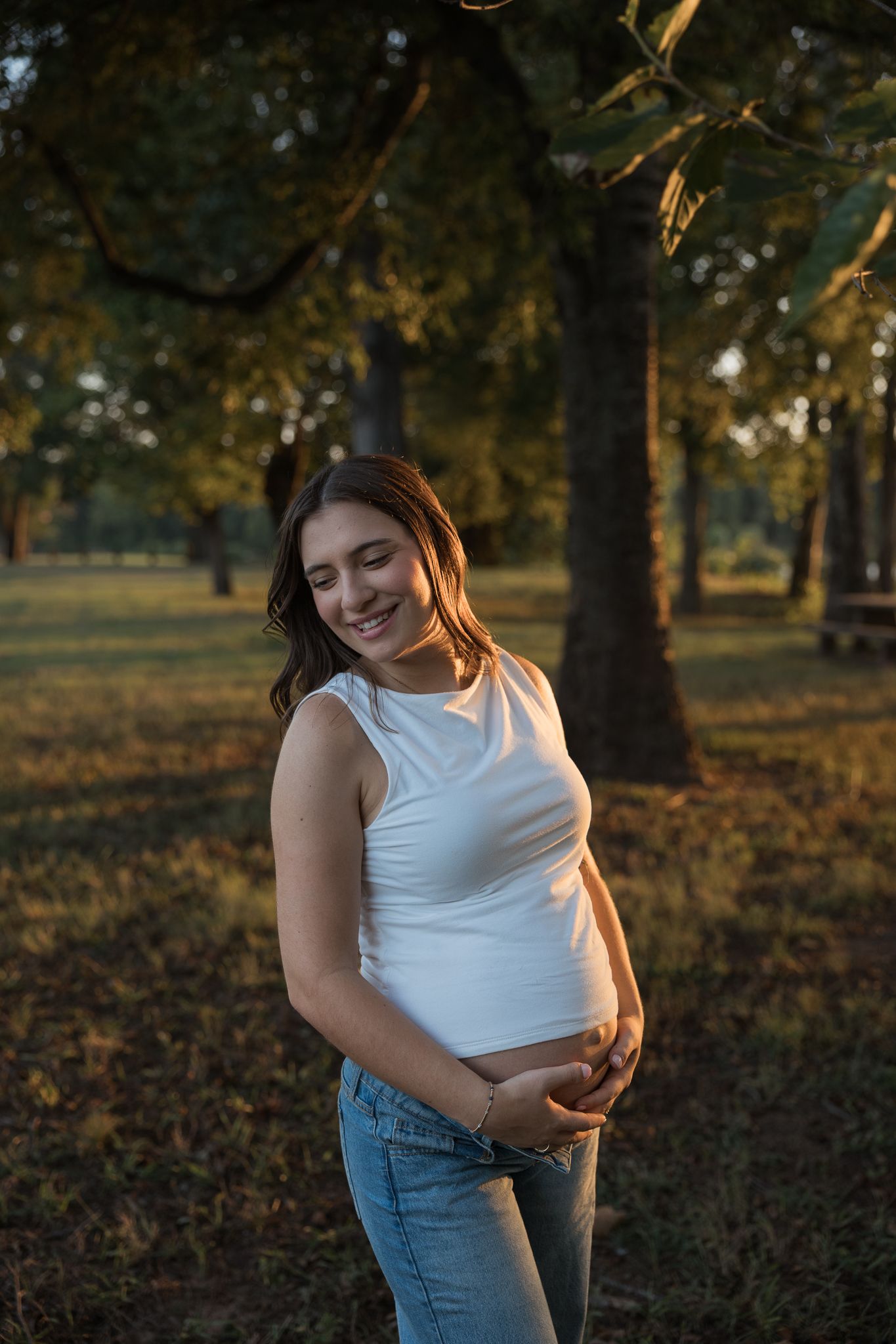 Glowing pregnant woman in white tank top smiling while holding baby bump in sunlit autumn park.