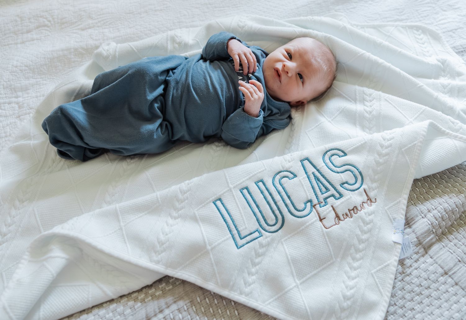 Newborn baby Lucas Edward in blue onesie lying on personalized embroidered white name blanket.