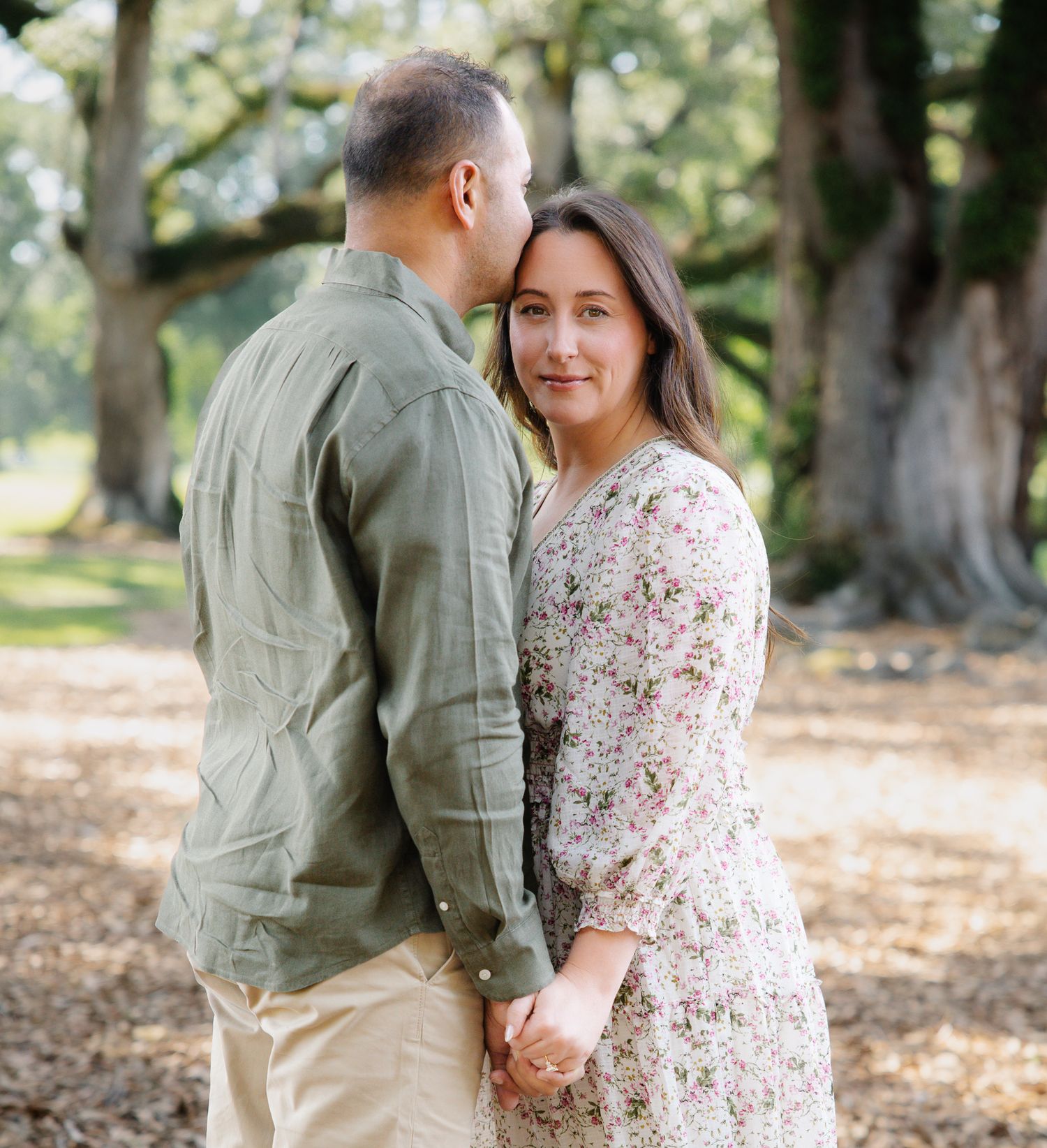 Couple holds hands and embraces outdoors among tall oak trees during a romantic engagement photo session.