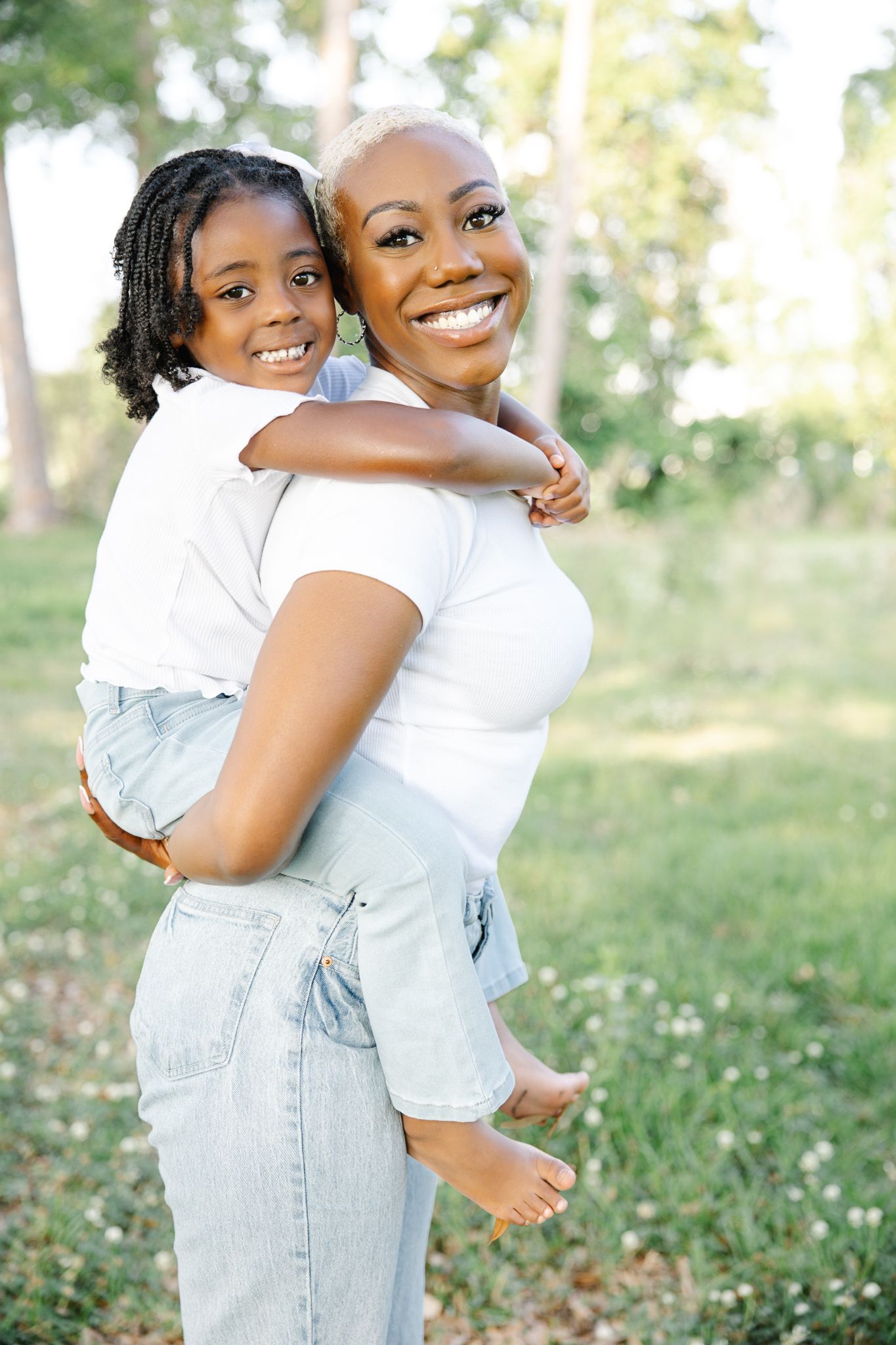 A joyful Black mother carries her smiling daughter piggyback in a sunny green park, both wearing white tops and denim.
