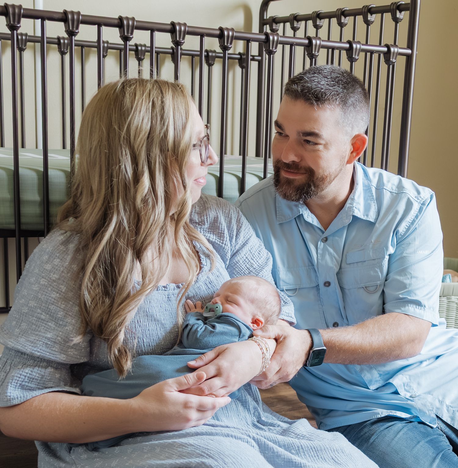 New parents sit together holding newborn baby boy in front of iron crib.