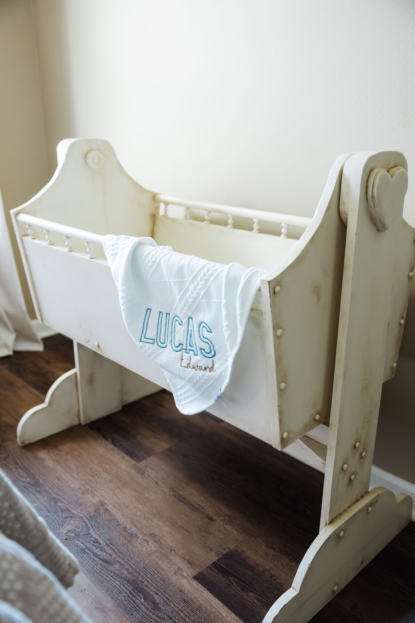 A white vintage wooden cradle with a personalized blue embroidered blanket reading Lucas Edward.