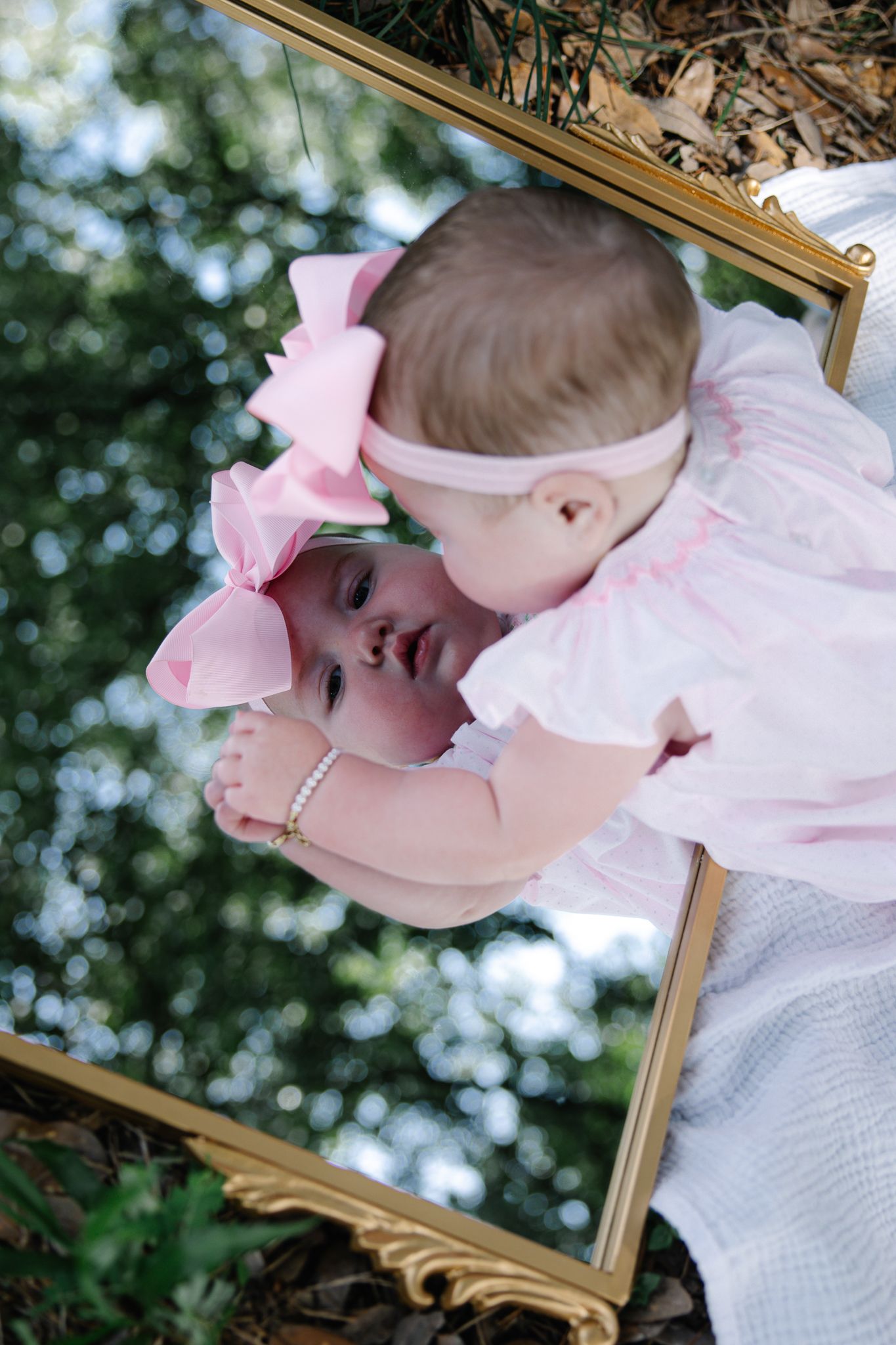 Adorable baby girl in pink bow headband looking at her reflection in a gold-framed mirror outdoors.