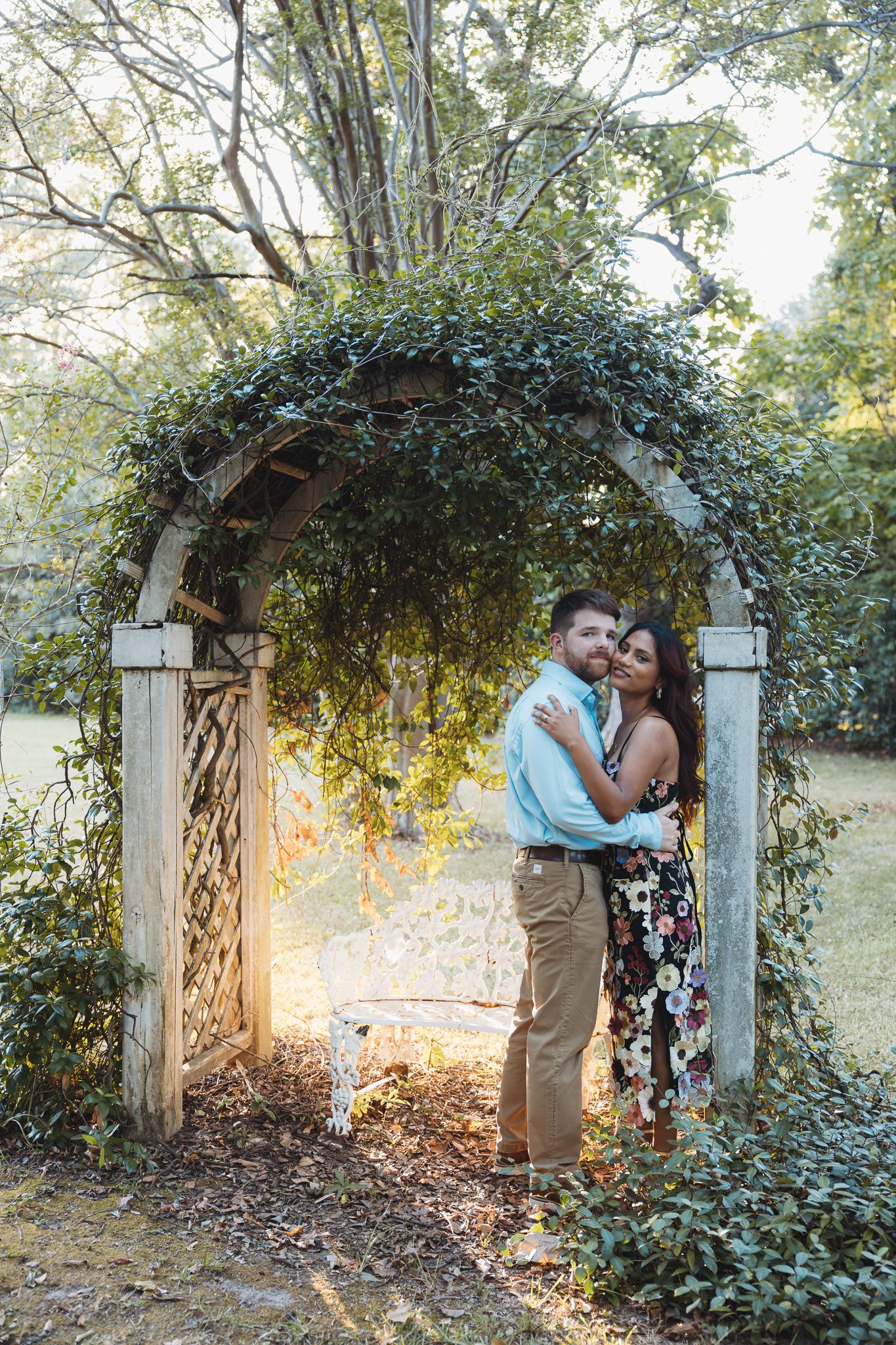 A couple embraces under an ivy-covered wooden garden arch in warm golden sunlight.