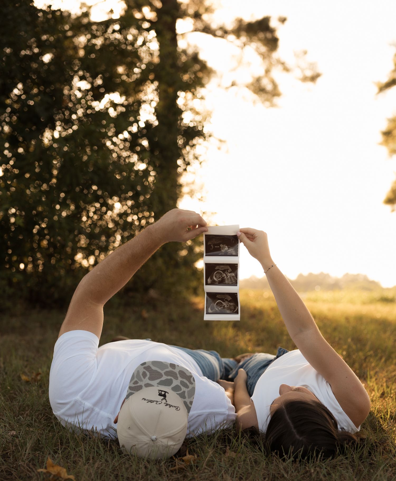 Couple lying on grass holding up ultrasound photos together in golden sunset light for pregnancy announcement.