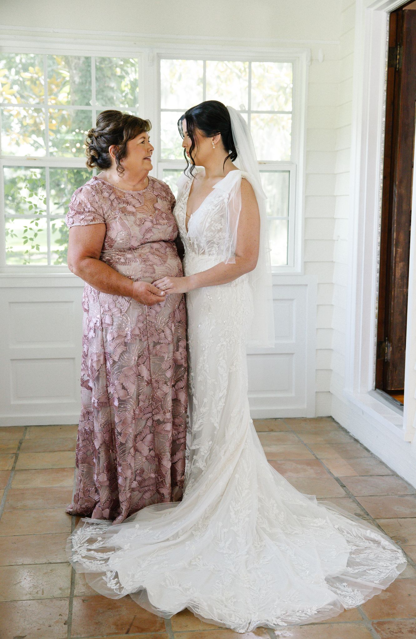 Bride in lace wedding gown shares an emotional moment with her mother in a bright white-windowed room.