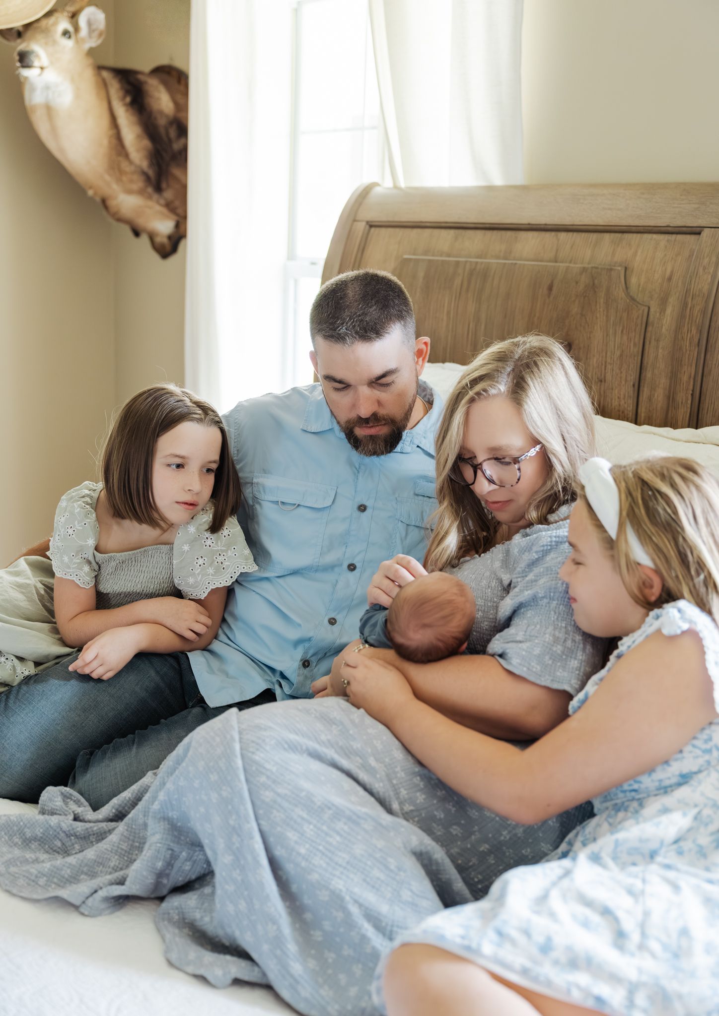 Father and three daughters sitting on bed together lovingly looking at their newborn baby boy.
