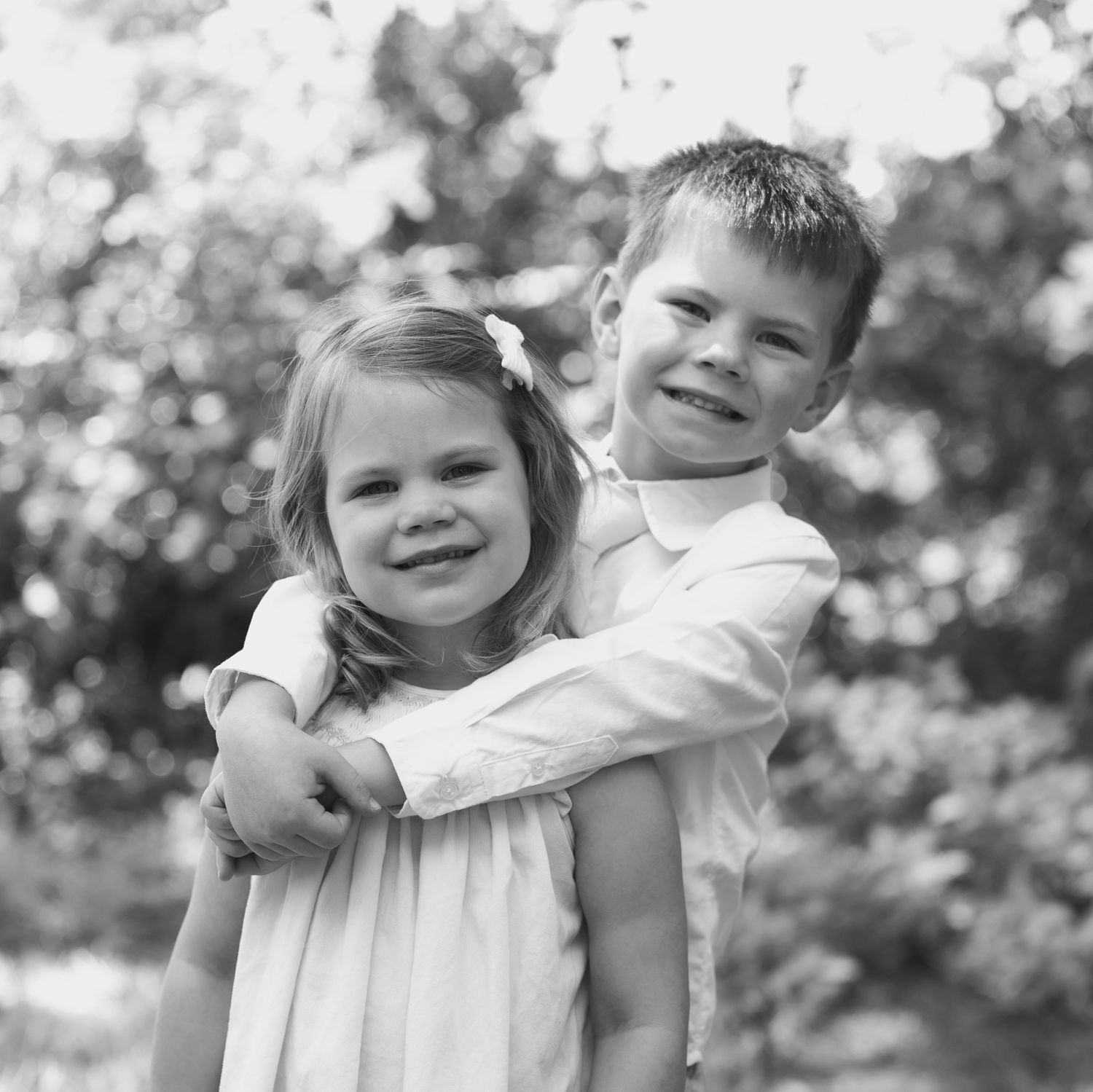 Black and white photo of two young siblings hugging and smiling outdoors among trees.