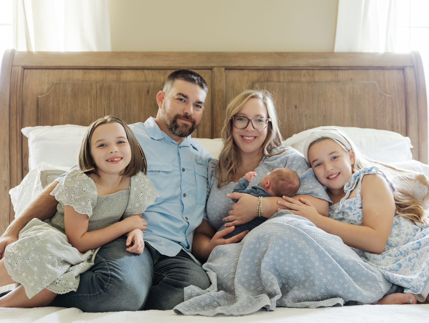 Happy family of five sitting on bed with newborn baby, two young daughters smiling together.