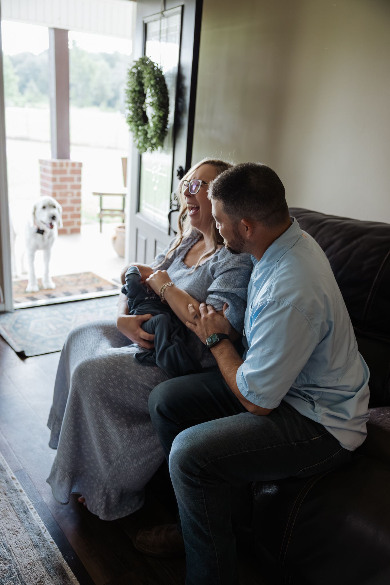 A couple sits on a sofa smiling at their newborn baby with a dog visible through the glass door.