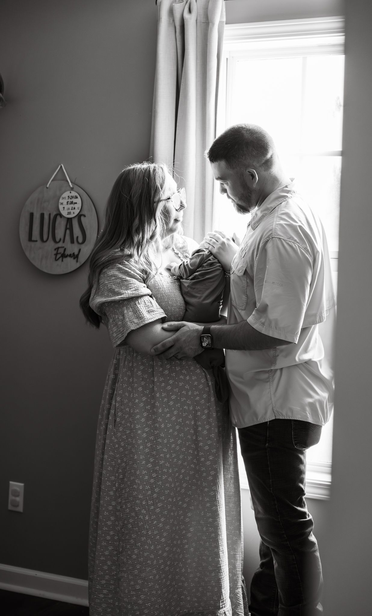 A couple stands by a window holding their newborn baby next to a Lucas name sign, black and white.