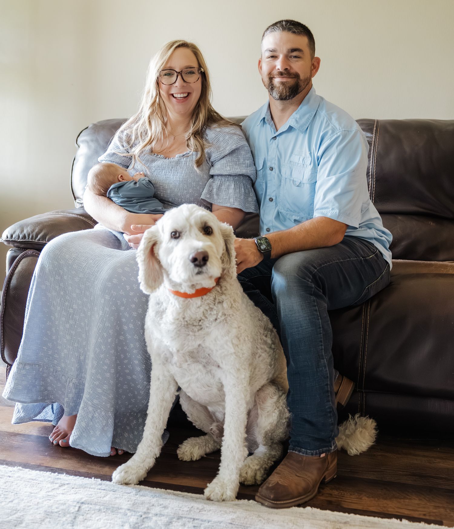 Happy couple with newborn baby and white goldendoodle dog sitting on brown leather sofa.