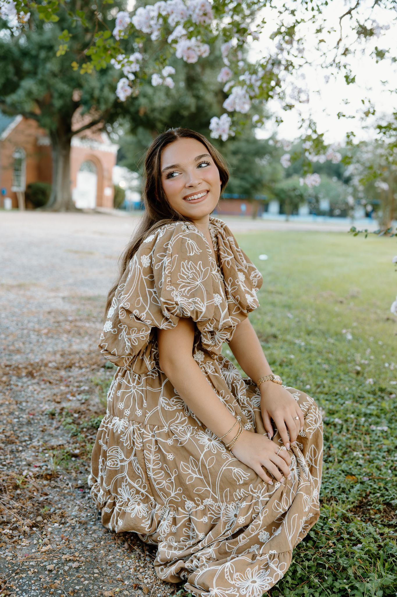 Young woman in floral tan dress sitting outdoors under blooming tree, smiling gracefully in a garden setting.
