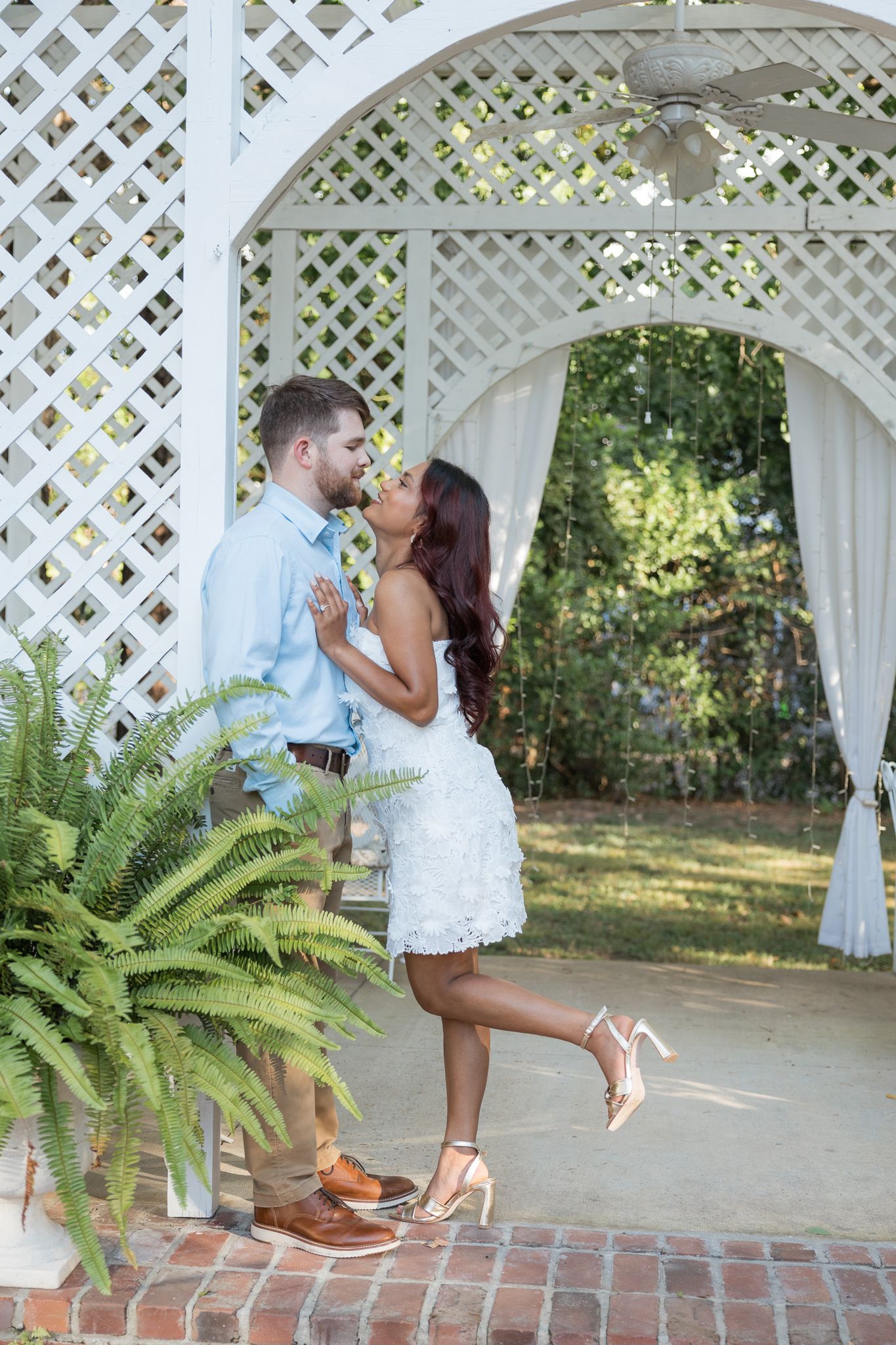 Couple sharing a romantic kiss under a white lattice gazebo, woman in white lace dress lifting her heel.