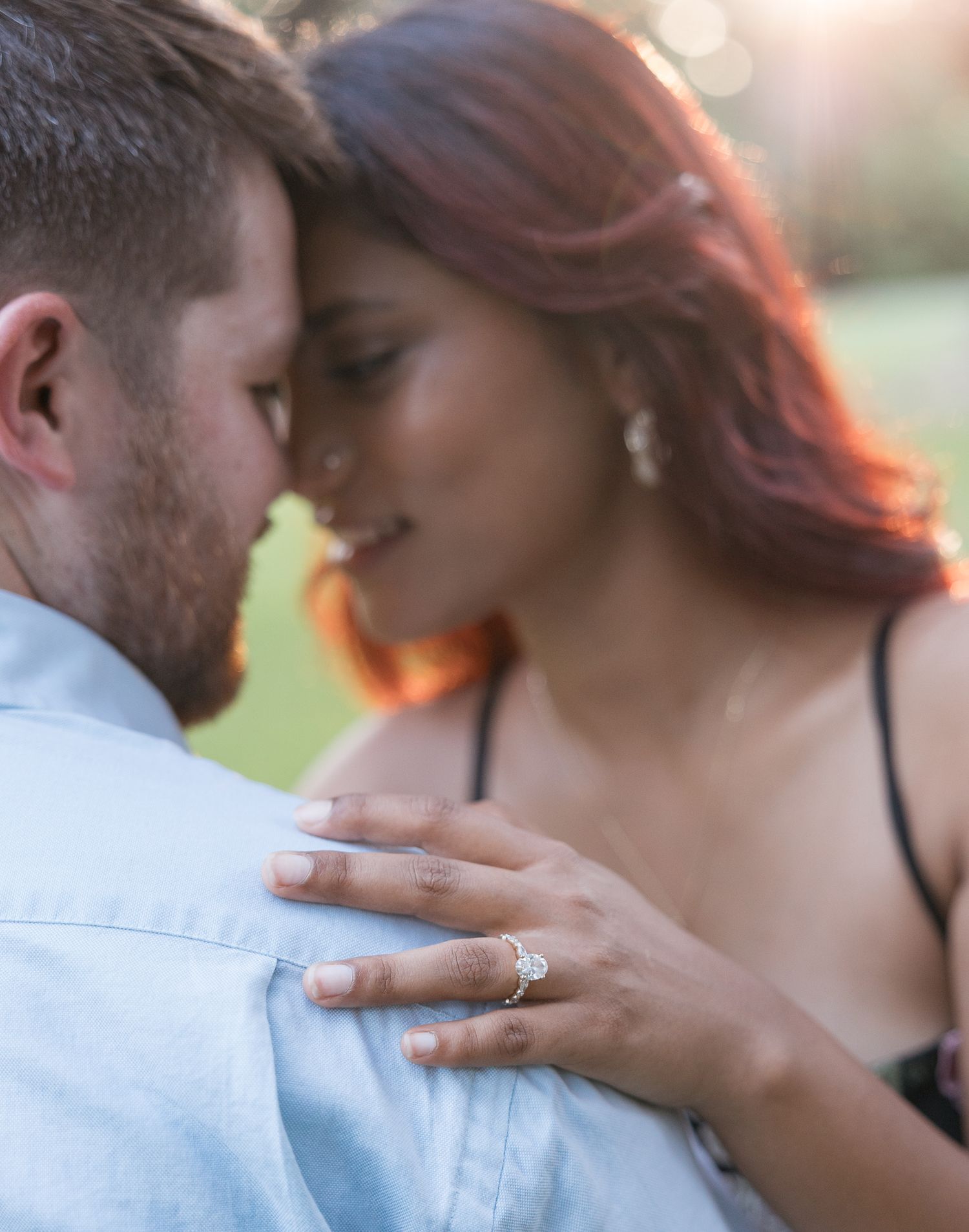 Couple sharing an intimate moment, woman with red hair wearing an engagement ring rests her hand on mans shoulder outdoors.
