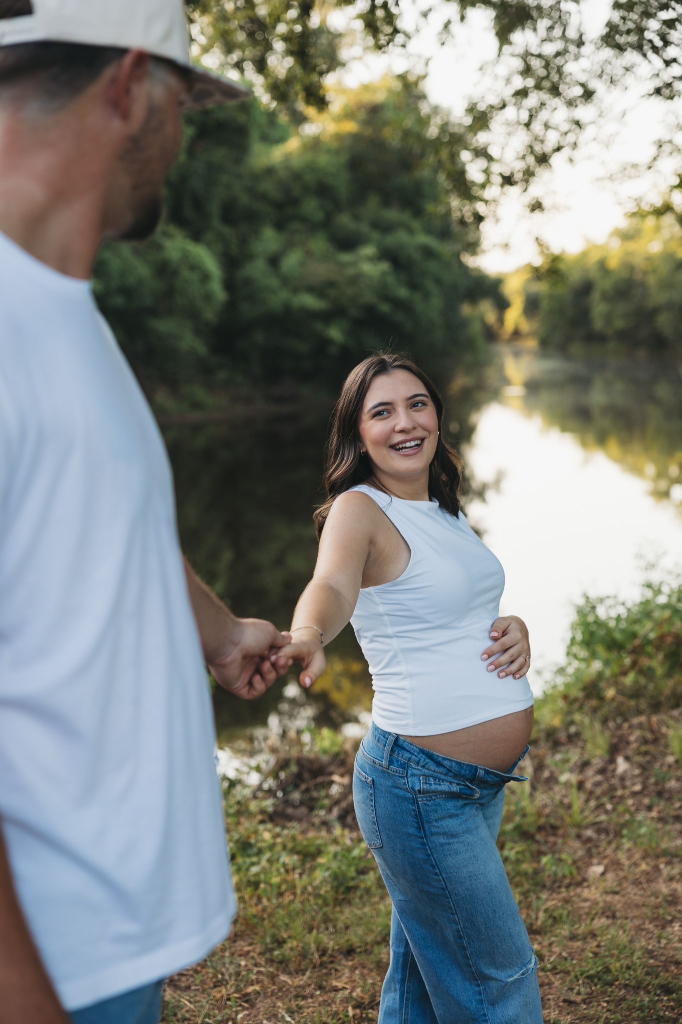 Smiling pregnant woman in white tank top and jeans holds hands with man near a serene river surrounded by lush greenery.