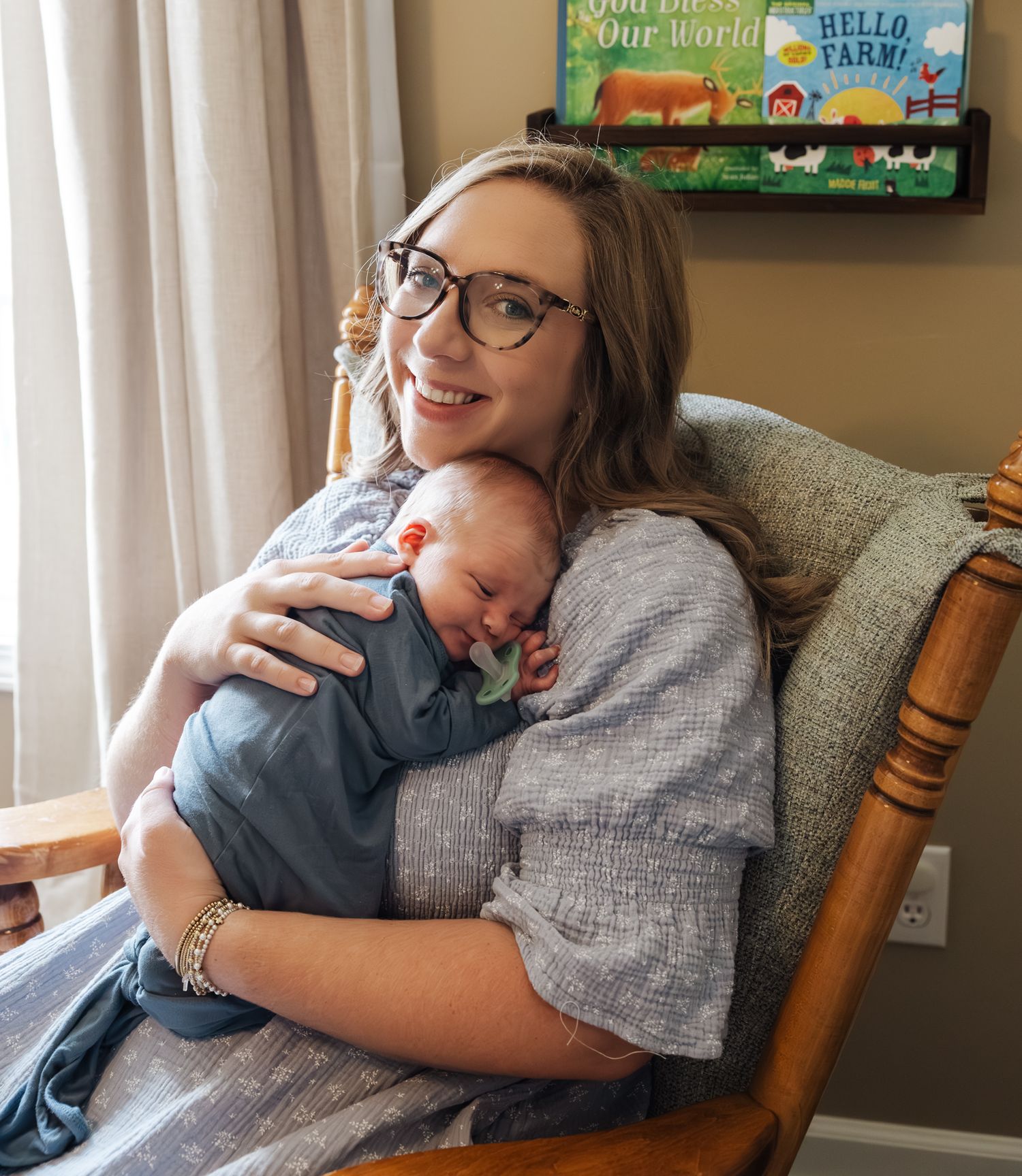 Smiling mom with glasses cuddles sleeping newborn in wooden rocking chair.