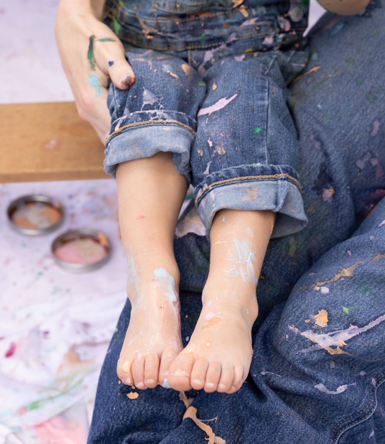 mother holding daughter in mommy and me photoshoot covered in paint and wearing denim overalls. close up of child's feet with paint in back