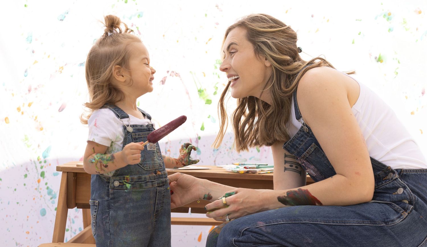 mother and daughter sharing laughter in mommy and me painting photoshoot, eating popsicle and sitting on mini picnic table covered in paint
